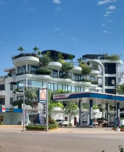 A high-end apartment building in Cambodia with a gas station next to it. It is four stories and has a rooftop with lots of green plants and small trees, and a cabana hangout area. The third floor rooms have balconies that are wavy-shaped, and have lots of green plants.