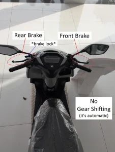 An image of an automatic motorbike. There is text with arrows pointing at the controls of an automatic motorbike. This image highlights the locations of the rear brake plus brake lock (on handlebar left-hand side) and front brake (on handlebar right-hand side). It also hightlights that there are no gear shifts, which a semi-automatic and fully manual motorbike would have. The brake lock is for parking on a hill.