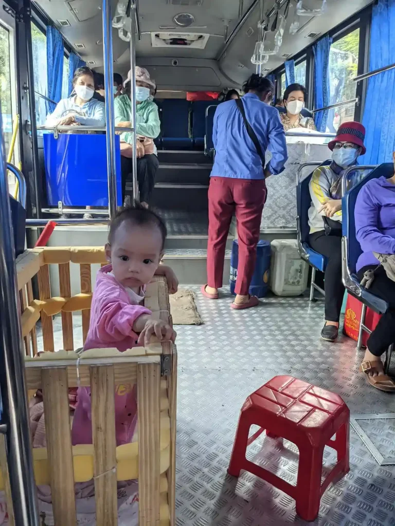 A public city bus in Vietnam. There is an onboard ticket seller, collecting fees. The seller has a makeshift play pen with their one year old child inside. The play pen is made of wood, and has padding zip-tied to it. The baby is standing up, while the bus is in motion, and is looking at the photographer of this image. There is a small, six inch tall, stool that the seller sits on next to the baby play pen.