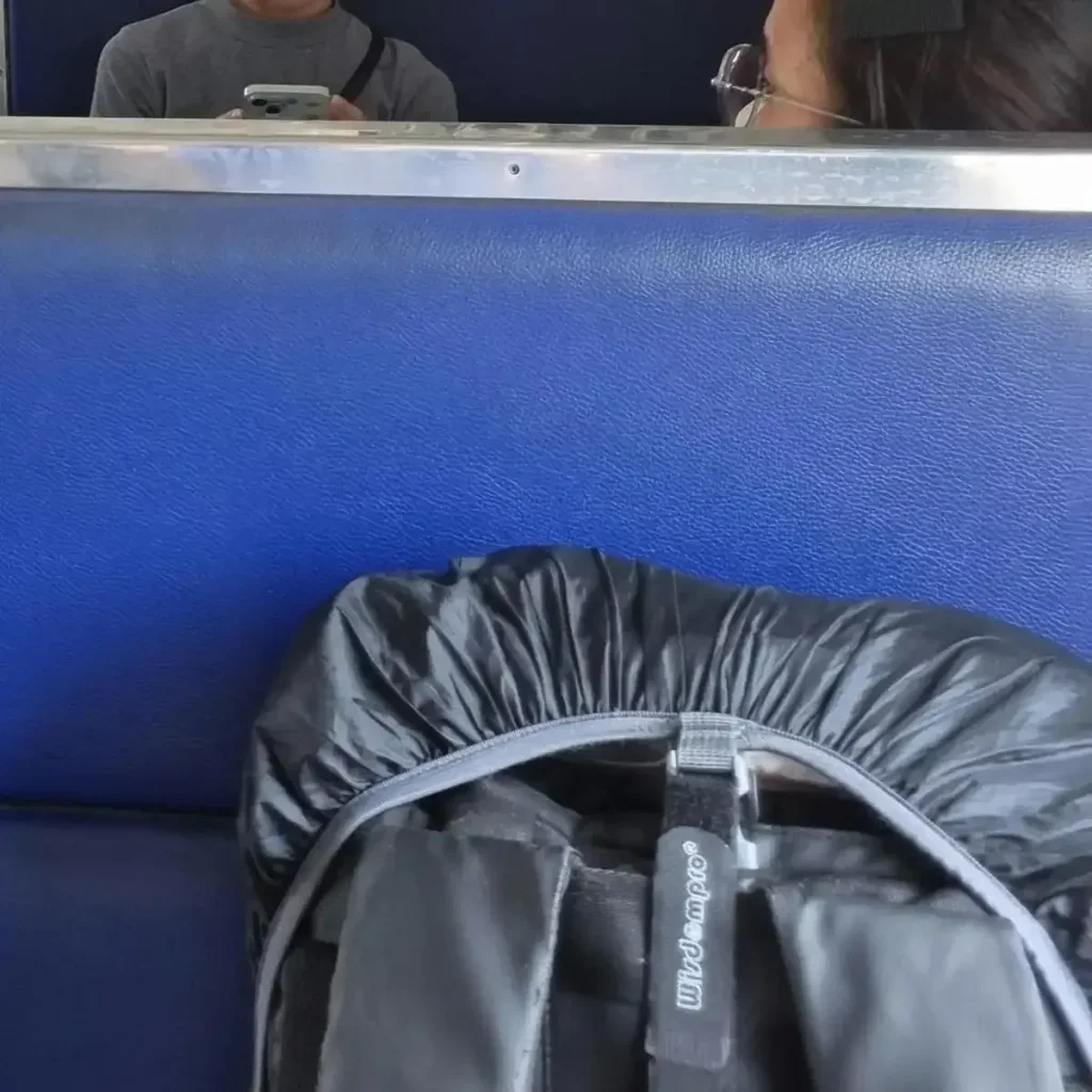 Carryon-sized 30 liter backpack (a.k.a in-cabin baggage) resting within a passenger's train seat. It has a rain cover over it that has an elastic band around it's permiter - good for stuffing things inside (between the bag and the rain cover), preventing theft. The rain cover, per the name, protects against water as it is made of water-resistant material. This picture shows a velcro compressor strap is used to securely strap the rain cover on, since the built-in straps & clips are not strong enough. Using the velcro strap allows the owner to use the cover as an extention - the built-in straps are often (but not always) not long enough when stuffing things inside.