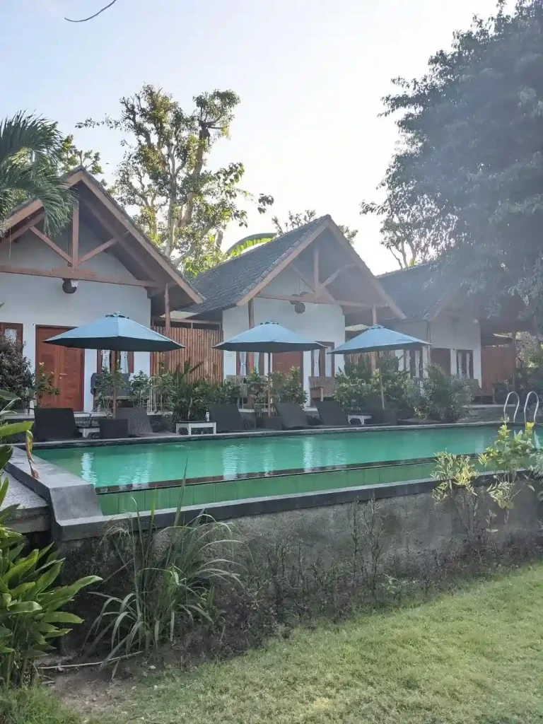 A hotel with three huts and a swimming pool outside. Several chairs and tables with umbrellas are along the pool. Trees and plants surround the area. The scenery is more about relaxing with nature.