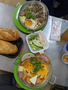 Two breakfast skillets with French bread on the side on a table in Vietnam with wet wipes as a napkin. This is called banh mi chao in Vietnamese. Different toping can be chosen. One of the dishes in this image have steak, onions, fried egg, cilantro, pate, and mayonaise. The other dish has ham, hot dog, cheese, fried eggs, cilantro, pate, and mayonaise. At the center is bowl of pickled vegetables, too.