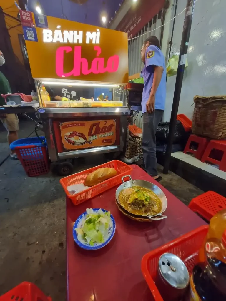 A small food stall specializing in a Vietnamese dish called banh mi chao in Saigon (aka Ho Chi Minh City), Vietnam. Banh mi chao, as pictured on a table next to the stall, is a skillet with a loaf of French bread. Although it is typically a breakfast dish, it is being served at night time in this image. The stall has a pleasant sign that lights up saying "Banh Mi Chao".