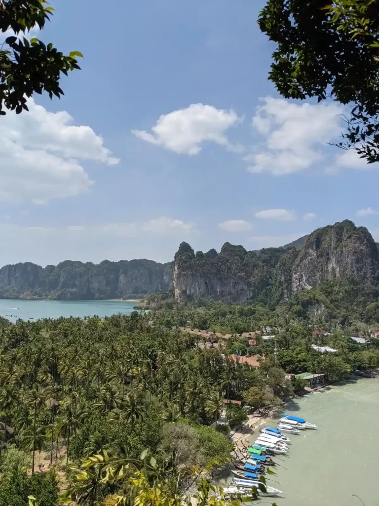 An overlook of two beaches with a stretch of land going between them, and steep mountains in the background. Palm trees cover the land, and boats are along one of the beaches.