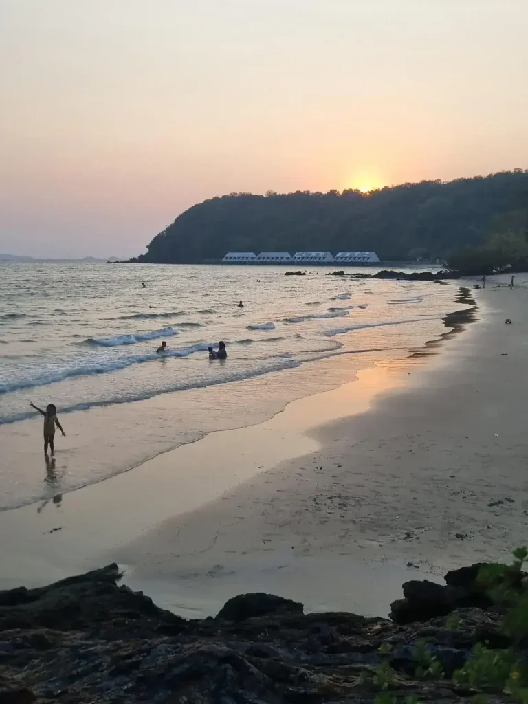 A beach at sunset time in Thailand. The photographer of this image is standing on an elevated rocky area looking at the beach with waves, and a small hilly area coverin the sun.