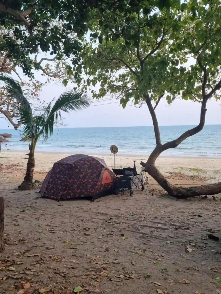 A camping tent setup next to two trees on a beach in Thailand, with no-one else in sight but the ocean. The camper also has some camping gear such as a wagon.