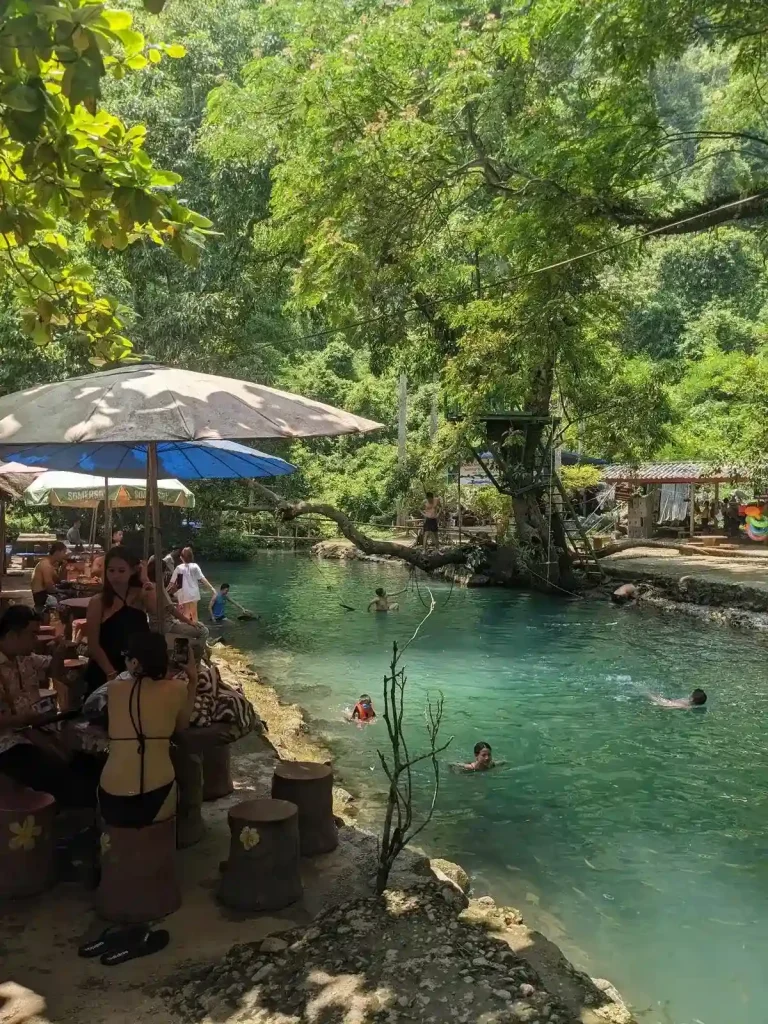 People at one of the Blue Lagoons in Vang Vien, Laos. Here, people are swimming in the lagoon, and having a picnic alongside it under the shade of umbrellas.