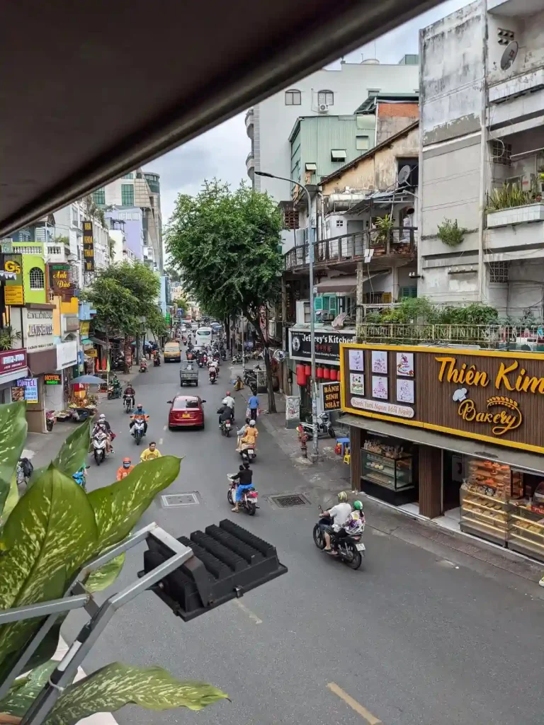 An image depicting busy big city life. The location is in Ho Chi Minh City (a.k.a. Saigon), Vietnam. The image is taken on the second story balcony overlooking a street with many motorbikes (a.k.a scooters), a very common mode transportation in Vietnam. Along the street is wall-to-wall shops, restaurants, and housing. This city has a very high population density, and there is little space between buildings, as they are typically connected together.
