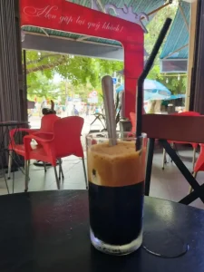 An image of a coffee on a table in a café in Vietnam. The coffee is: Café (coffee) den (black) da (iced) phin (i.e. made with a phin brewing filter instead of using espresso).
