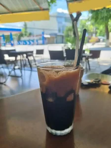 An image of a coffee on a table in a café in Vietnam. The coffee is: Café (coffee) den (black) da (iced) phin (i.e. made with a phin brewing filter instead of using espresso).
