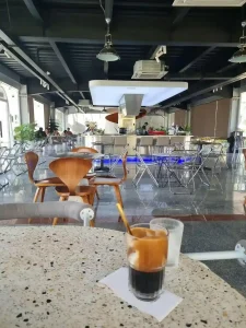 A café in Vietnam. The image shoes an iced black coffee in a glass, Vietnamese style, on the table. The coffee has some sugar inside that has been whisked within the coffee, and therefore has a coffee foam on the upper part of the glass