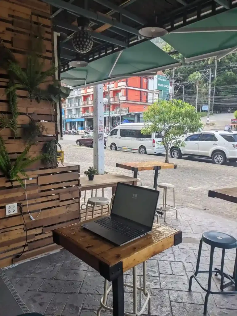 A workspace for remote working at a café. A remote worker is setup in a café with open-air outdoor seating. Their laptop is on the table with bar stool seating.