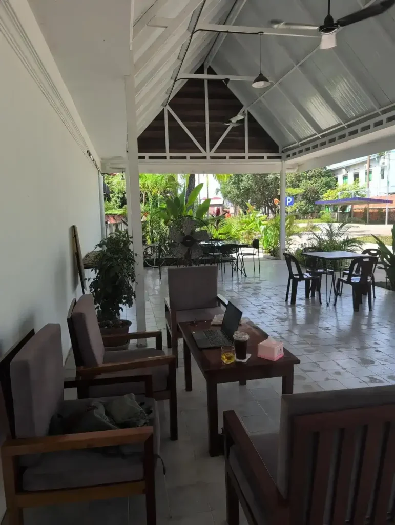 A workspace for remote working at a café. A remote worker is setup in a café with open-air outdoor seating. Their laptop is on the table, and they have comfortable seating. A fan is near the table to keep them (and their laptop) cool.