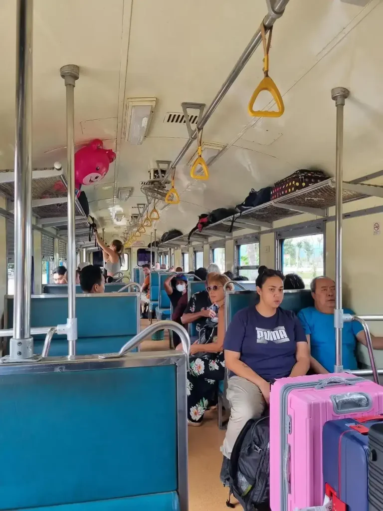 Bench seating on the inside of the Cambodian train cabin. Each bench seat can seat 2 adults. The bench seating is in groups of 2 bench seats for 4 adults total, one bench seat facing forward plus another facing rearward. There is a little bit of leg space in between. The layout arrangement is bench seating on the left-hand side, then a middle row ailse, and another set of bench seats on the right-hand side. Windows slide down for a cool breeze. There is a luggage rack mounted above all of the seats. Safety handles are mounted on the ceiling within the ailse.
