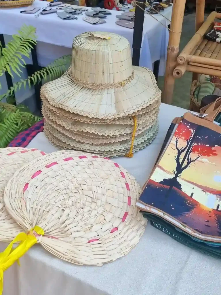 A table of artisan souvenirs in Cambodia; including a straw hat, hand-waved air fan, and tote bags with various motifs printed on them of local surrounding such as from within Angkor Watt Park.