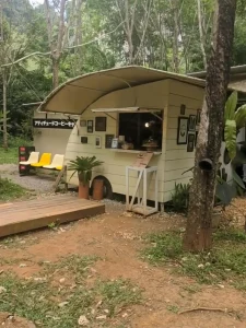 A camping-themed café with tents within a rubber tree farm in Krabi, Thailand. The image shows the ordering counter that is shaped like a small camper (i.e. the kind that is towed behind a vehicle in the USA).