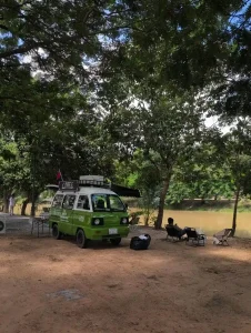 A camping-themed café with a small mini van used as the ordering counter and tools inside to brew coffee. It is parked along a river with seating.