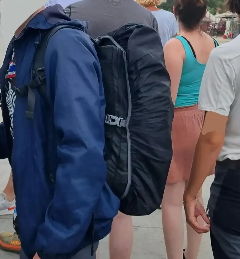 Passenger wearing a 30 liter carry-on backpack with a rain cover stretched over it. The rain cover protects against rain and prevents theft by anyone reaching into the bag. And, since this rain cover has an elastic band around it's perimeter, the bag owner can stuff things inside, between the bag and rain cover. This acts as an extention of the bag, and/or a way to quickly access access things while on the go, such as a spare t-shirt and shorts for sleeping while traveling.