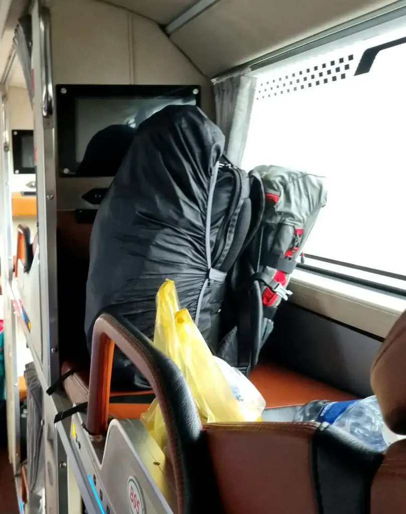Two carryon backpacks (a.k.a in-cabin baggage) resting within a passenger's bus seat. The seat is actually a bunk bed on a sleeper bus in Southeast Asia. One bag is the main backpack (medium sized 30 liter bag), and it has a rain cover over it. The other bag is a smaller 18 liter day bag that is not full. This small bag has two velcro strap wrapped around it to compress the bag so that it takes up less space.