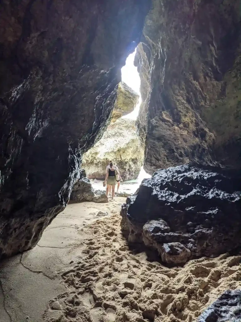 The opening of a sandy cave that two people are walking out of to the ocean.