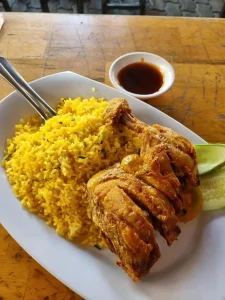 A plate of Com Ga, which is Vietnamese meaning "Rice Chicken". The rice is fried, the chicken deep fried, and there is a side of brown sauce and sliced cucumber.