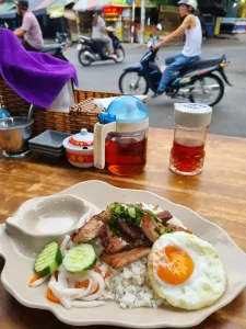 A dish of pork rib on rice with a fried egg, pickled vegetables, and sauce on the side. The table is on a street corner watching motorbike (aka scooter) traffic riding by. These restaurants serve food called "Com Tam" (gum tam), meaning "broken rice", which refers to the type of rice served - it is small as if it was broken in half.