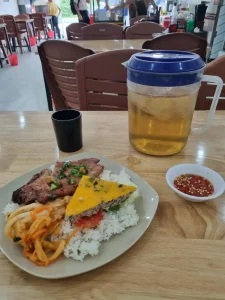 A dish of pork rib on rice with a Vietnamese meat loaf, Korean kimchi instead of pickled vegetables, and spicy-looking soy-based sauce on the side. A jug of iced team is on the table for customers. The table is inside a Com Tam restaurant. These restaurants serve food called "Com Tam" (gum tam), meaning "broken rice", which refers to the type of rice served - it is small as if it was broken in half.