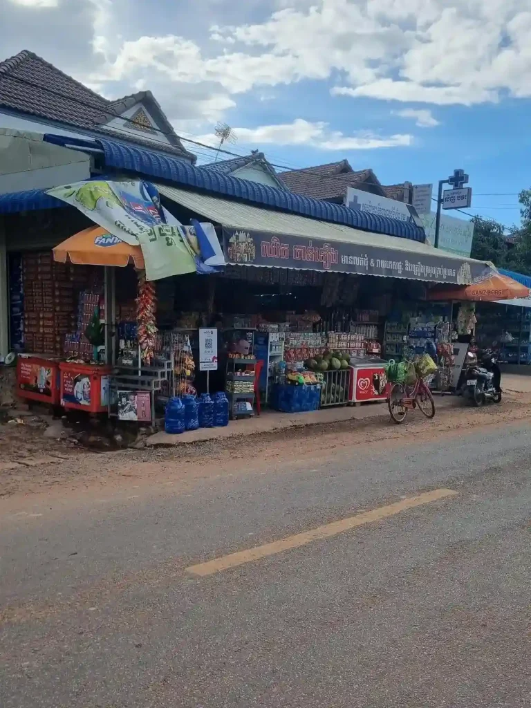 A local family-owned "mom and pop" convenience store in Cambodia extending over the entire sidewalk. This is very common all throughout Cambodia and Southeast Asia, in general. Located just right off the street, the store does not have a wall, making it is easy to stop and walk right into the store. Inside, the store has twenty liter water jugs, lots of snacks, a cooler with ice, a deep freezer with ice cream, and a cage with coconuts inside and ontop of it. A corrugated sheet metal roof covers the store. But since the store extends completely over the sidewalk, a canvas awning has been added to cover this extension.