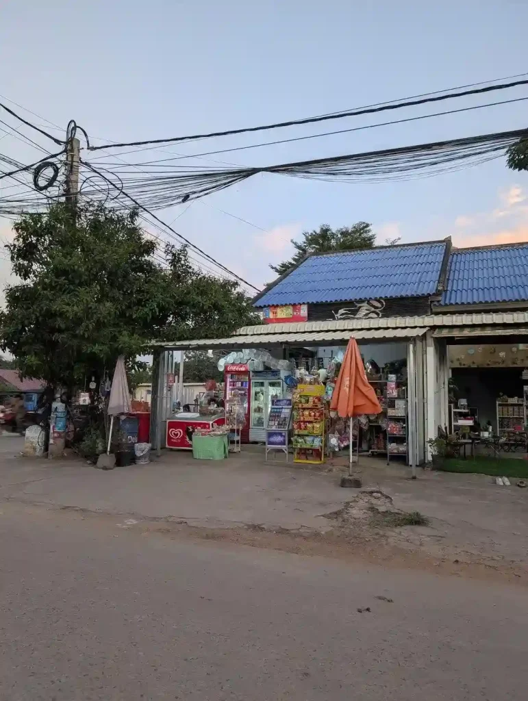 A local family-owned "mom and pop" convenience store in Cambodia. This is very common all throughout Cambodia and Southeast Asia, in general. Located on a street corner, the store has not walls on the two sides that face the street. Inside, the store has two refrigerators with drinks, a deep freezer with ice cream, and racks with various snack food on them. A corrugated sheet metal roof covers the store.