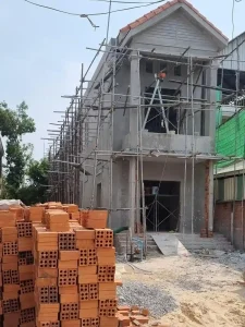 An image showing the construction method in Southeast Asia for building homes. This two story house is still under construction. A concrete skeleton was constructed on a foundation. Then, brick was laid to fill in the skeleton, building the outer and inner walls of the home. Finally, concrete is smeared over the brick and smoothed out for a smooth texture. Roofing is made of clay. A stack of clay bricks are in front of the house.