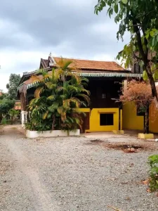 A traditional wooden house in Southeast Asia. The home is a stilt house, meaning it is elevated off of the ground. However, the bottom section, which traditionally is just for storage, has been converted to a living space. The bottom has had walls constructed using brick and concrete. A front door was also added to the bottom converted half. The gravel makes up the entire driveway and front lawn area.
