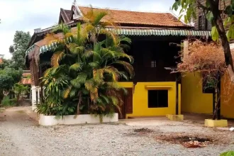 A traditional wooden house in Southeast Asia. The home is a stilt house, meaning it is elevated off of the ground. However, the bottom section, which traditionally is just for storage, has been converted to a living space. The bottom has had walls constructed using brick and concrete. A front door was also added to the bottom converted half. The gravel makes up the entire driveway and front lawn area.