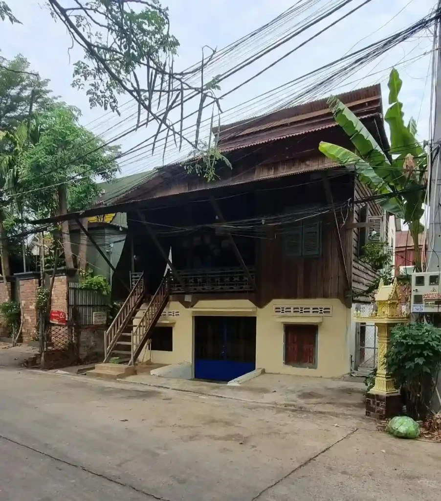 A traditional wooden house in Southeast Asia. The home is a stilt house, meaning it is elevated off of the ground. However, the bottom section, which traditionally is just for storage, has been converted to a living space. The bottom has had walls constructed using brick and concrete. The front porch on the traditional upper half has a covering and wooden stairs to go outside.