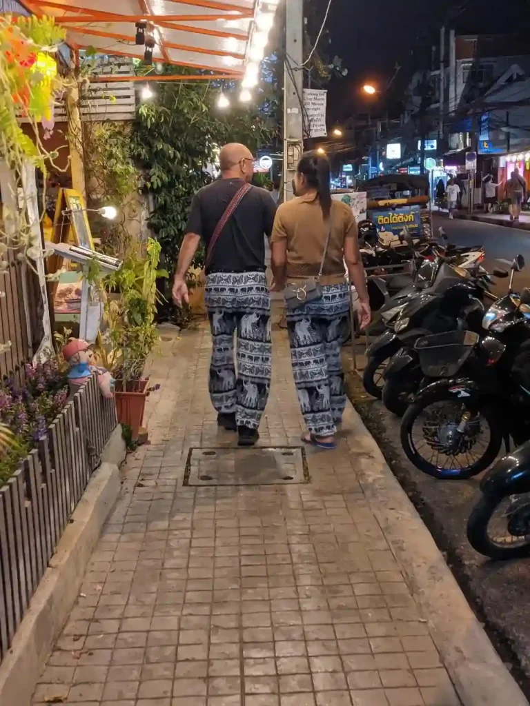 A couple walking together down the street in Thailand wearing chang pants. Chang means elephant in Thai, so these pants have imprints of elephants on them. These pants are the quintessential tourist clothing.