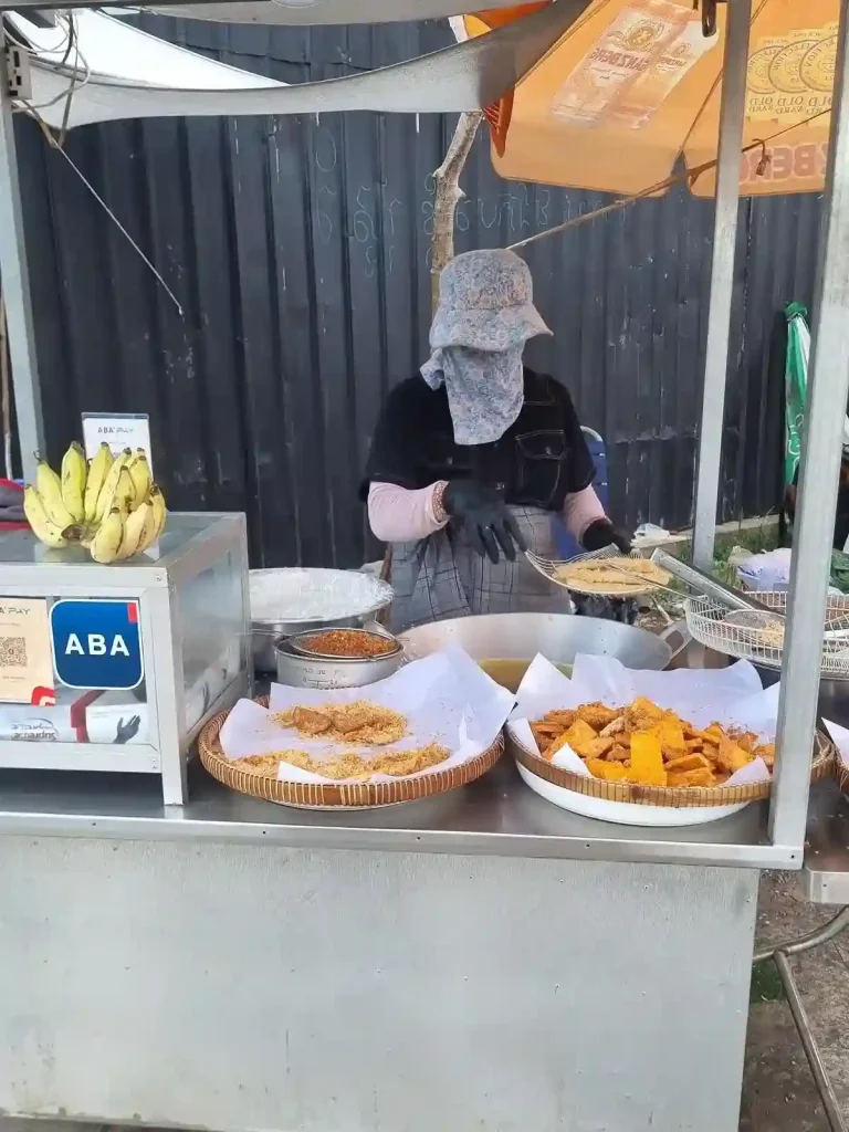 A food seller's kiosk along a road in Cambodia, in which they are preparing and have a display of deep fried bananas in a baskets for sale. This is a common snack throughout Southeast Asia. Bananas are peeled, sliced, covered in a battered, and then deep fried.