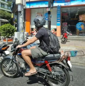 A Detech Honda Win motorcycle that is fully manual (uses a hand clutch lever, in part, to change gears). The rider is a foreigner from a Western country, riding in a city in Vietnam. This motorbike has a home-made rack on the back to mount baggage during a motorbike adventure.