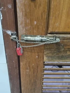 An image of how to lock a door with a small-link metal chain and travel lock. The chain is small enough to feed through holes in the door latch shown, mounted on the door. It also feeds through the latch mounted on the door frame on the wall. This ties the door to the door frame. A small TSA four pin travel lock connects & locks both ends of the chain together. This means that the door cannot be opened until the lock & chain are taken off.
