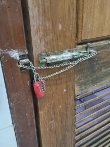 An image of how to lock a door with a small-link metal chain and travel lock. The chain is small enough to feed through holes in the door latch shown, mounted on the door. It also feeds through the latch mounted on the door frame on the wall. This ties the door to the door frame. A small TSA four pin travel lock connects & locks both ends of the chain together. This means that the door cannot be opened until the lock & chain are taken off.