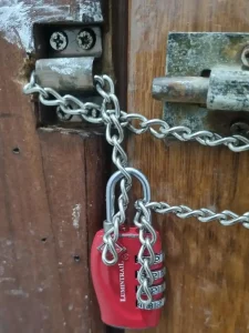 A close-up of a small-linked chain that is fed through the latch mounted on the door frame on the wall. A small TSA four pin travel lock connects & locks both ends of the chain together. Not show is how the chain also is fed through a hole in a door latch mounted on the door. This ties the door to the door frame. This means that the door cannot be opened until the lock & chain are taken off.