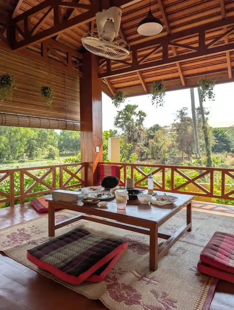 A large hut in Cambodia for eating in with a table and floor seating consisting of a mat and cushions. A rural view can be seen from within the hut. Air fans are mounted for a cool breeze, and bamboo curtains are rolled down to provide shade and block the sun.