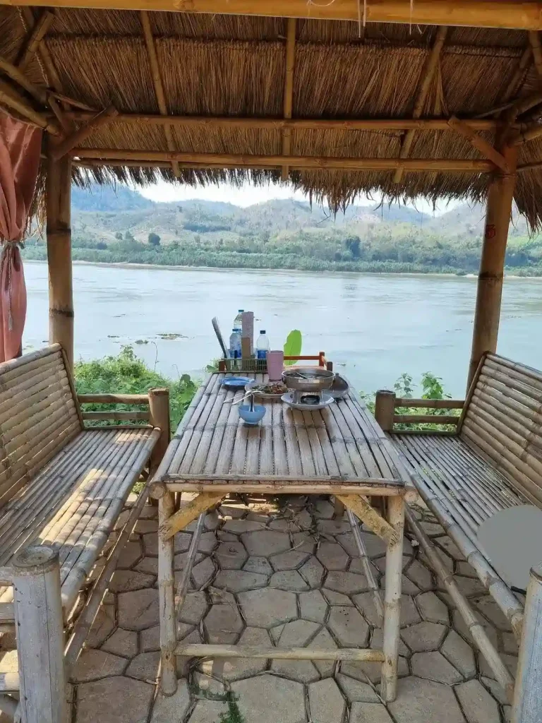 A small hut with bench seating and a table at an outdoor restaurant in Thailand. This is located along the Mekong river in rural Thailand. A couple of palm trees are on the property.