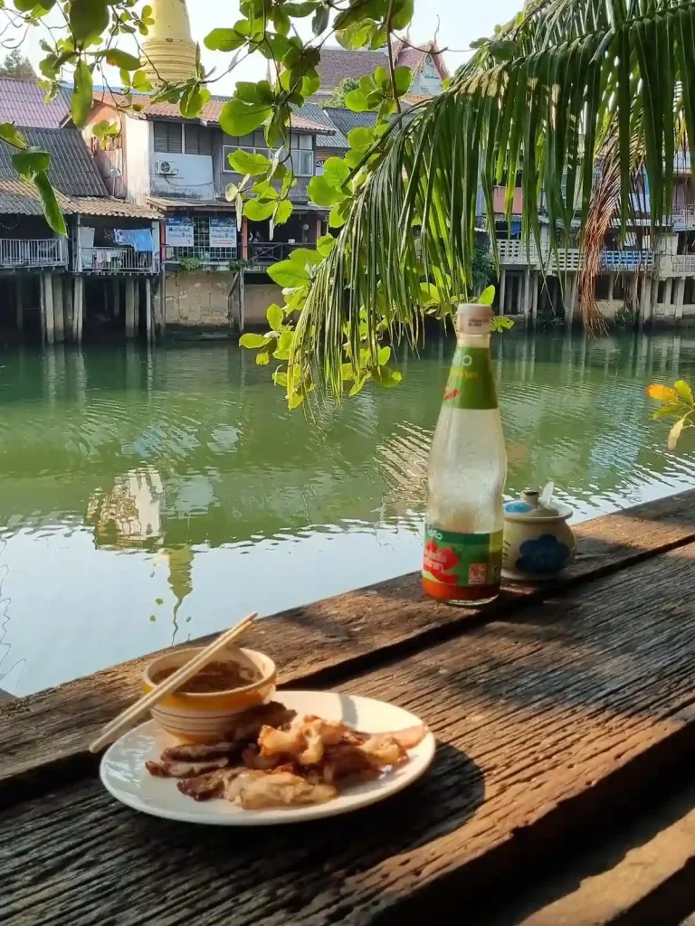 A prepared Thai food dish placed on a table. The eating area is a deck that overlaps onto a river with a golden temple in the background. There is also a line of housing, tightly spaced, that are also overlapping onto the river. The river is not very wide, you could throw a stone across, just barely.