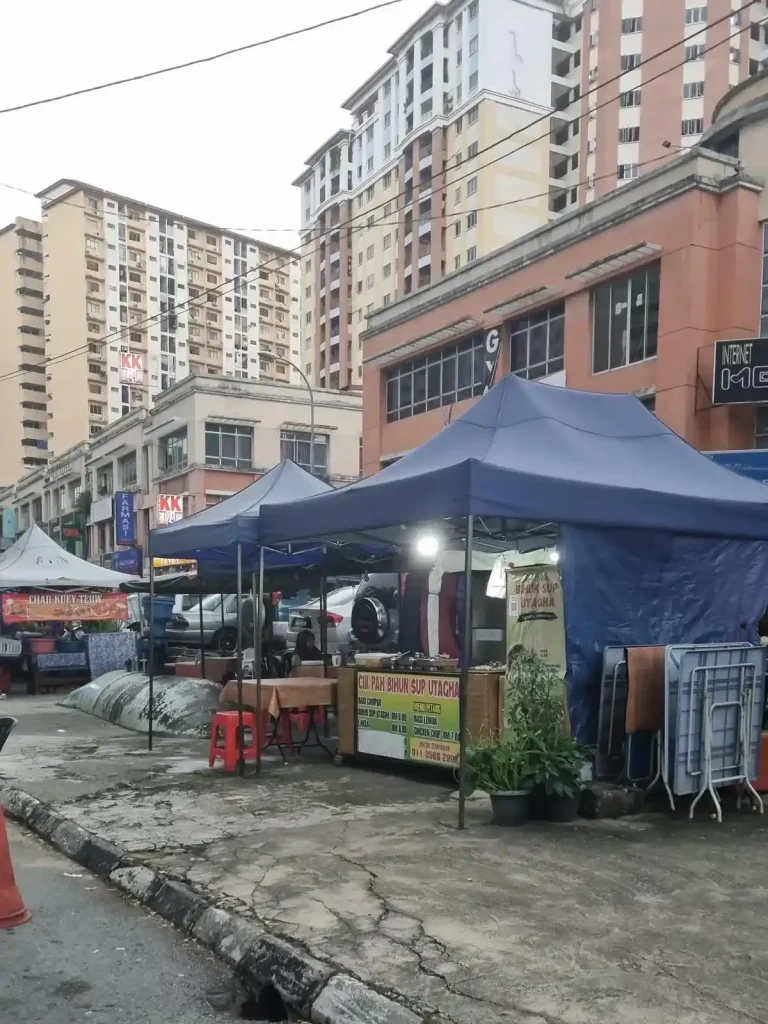 A food stall on the sidewalk of a street in Kuala Lumpur, Malaysia with high rise housing an and another food stall with canopy in the background. There are two pop-up canopies and a tarp covering the area with seating, the food stall, and a kitchen area underneath.
