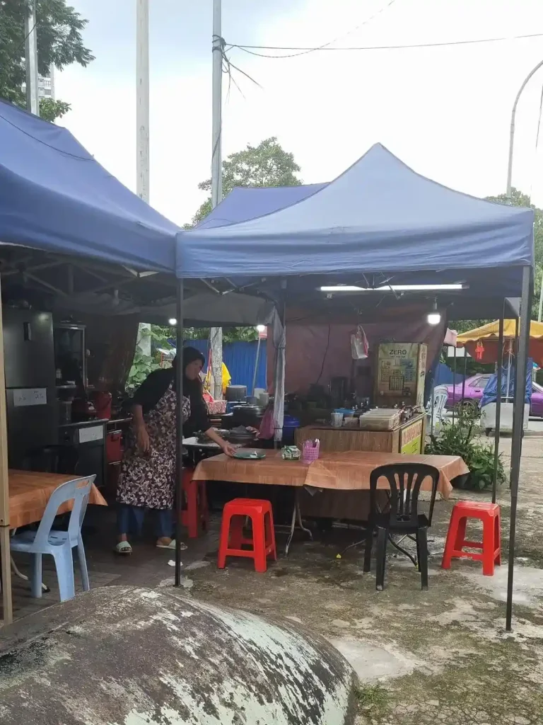 A food stall on the sidewalk of a street in Kuala Lumpur, Malaysia. There are two pop-up canopies and a tarp covering the area with seating, the food stall, and a kitchen area underneath. A full-sized refigerator and a stove are visible.