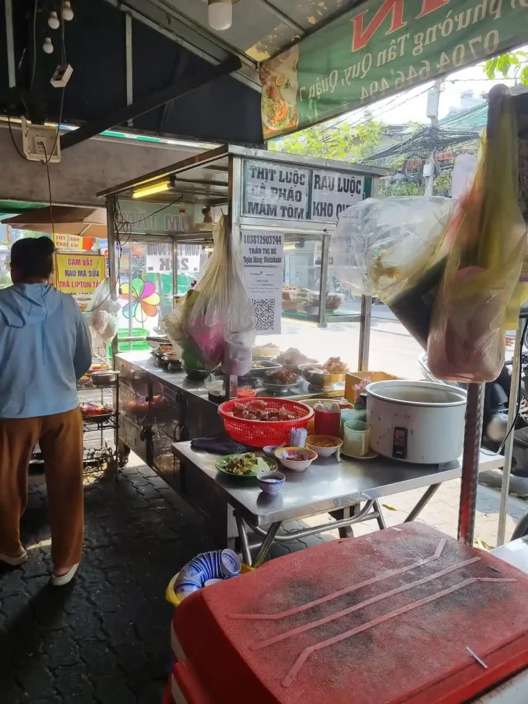 A food stall on the sidewalk of a street in Vietnam. An overhead awning covers the sidewalk, and thus the food stall.