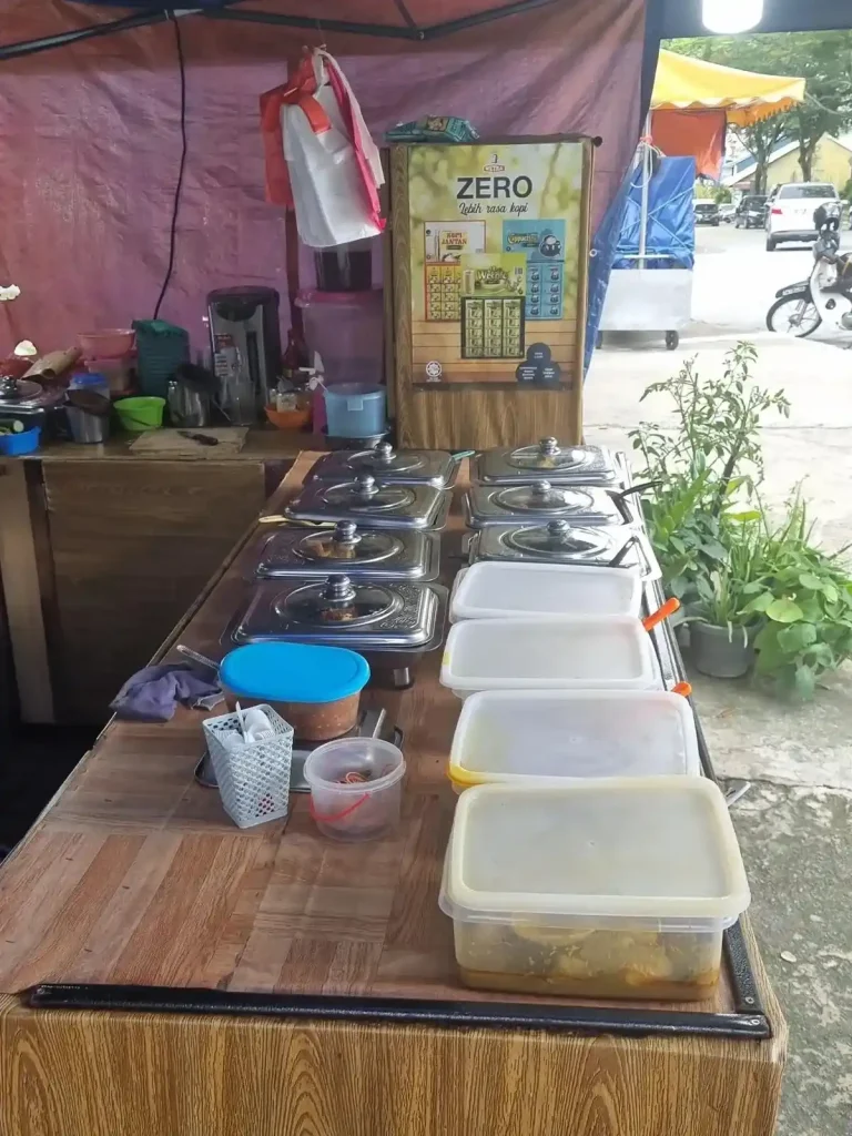 Food on display on a table for customers to choose from at food stall on the sidewalk of a street in Kuala Lumpur, Malaysia. Four dishes are in tupperware and seven are in metal containers with a heating element underneath.
