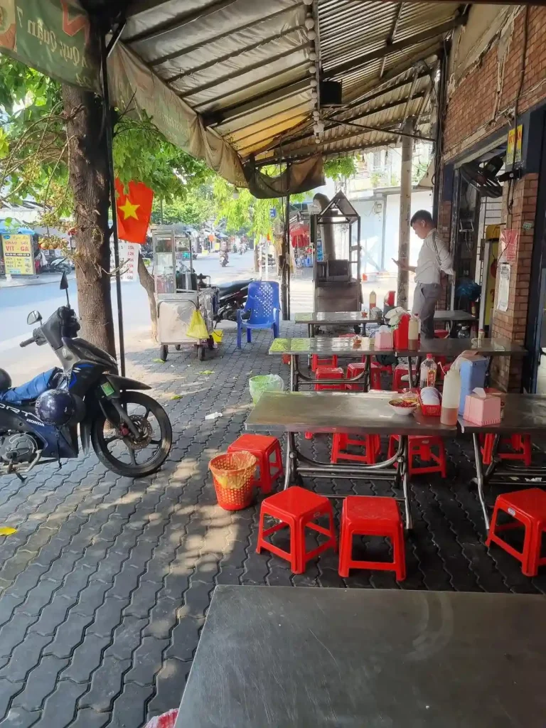 The seating area of a food stall on the sidewalk of a street in Vietnam. An overhead awning covers the sidewalk, and thus the seating area. A motorbike (scooter) is parked on the sidewalk next to one of the tables. It is common in Vietnam to use sidewalks as a parking lot, and some busy businesses have a security guard that will tend the motorbikes.