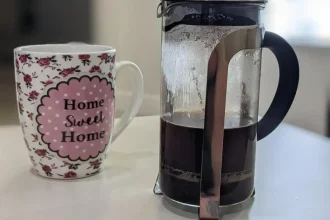 A coffee mug and a French press for brewing coffee on a table in an apartment. The coffee mug says "Home Sweet Home" on it. A french press has a glass tube (resembles a long cup), a plunger filter, handle, and a lid with an opening. Ground-up coffee beans are put inside the glass tube, hot water is added to brew the coffee, then the plunger attachment is put onto the glass tube. This attachment has a lid with a plunger going through it. The lid has an opening to pour the coffee into a coffee mug. After the coffee has finished breweing, the plunger is pushed downward throught the glass tube. It has a metal mesh filter on the end of it that pushes the ground coffee to the bottom of the glass tube, so that it does not get poured into a coffee mug. Drinking the grounds is not ideal. Therefore, the coffee grounds at the bottom are separated from the liquid coffee.
