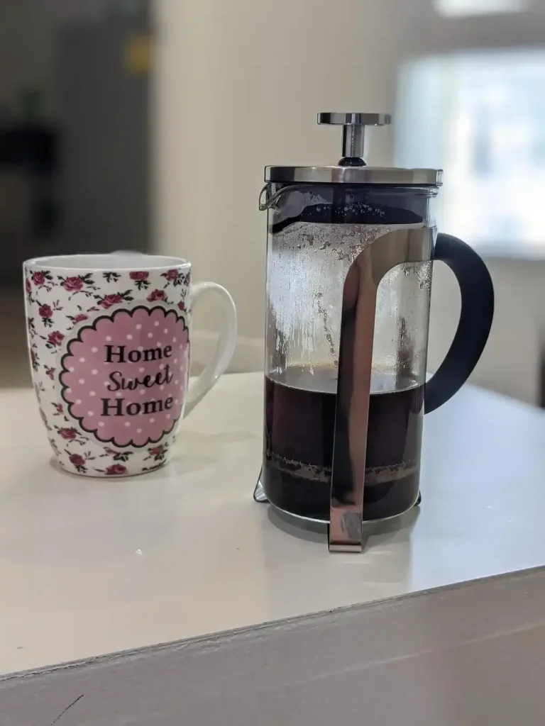 A coffee mug and a French press for brewing coffee on a table in an apartment. The coffee mug says "Home Sweet Home" on it. A french press has a glass tube (resembles a long cup), a plunger filter, handle, and a lid with an opening. Ground-up coffee beans are put inside the glass tube, hot water is added to brew the coffee, then the plunger attachment is put onto the glass tube. This attachment has a lid with a plunger going through it. The lid has an opening to pour the coffee into a coffee mug. After the coffee has finished breweing, the plunger is pushed downward throught the glass tube. It has a metal mesh filter on the end of it that pushes the ground coffee to the bottom of the glass tube, so that it does not get poured into a coffee mug. Drinking the grounds is not ideal. Therefore, the coffee grounds at the bottom are separated from the liquid coffee.