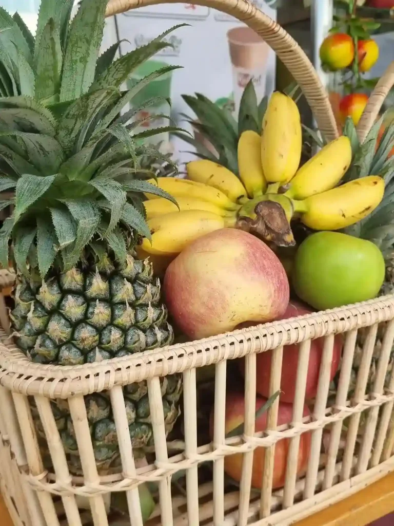 A basket of fruit at a café in Southeast Asia, which is used to make fruit smoothies. Inside is a pineaple, three red apples, one green apple, and a set of eight bananas. The bananas are a strain that are small, about the length of a finger.